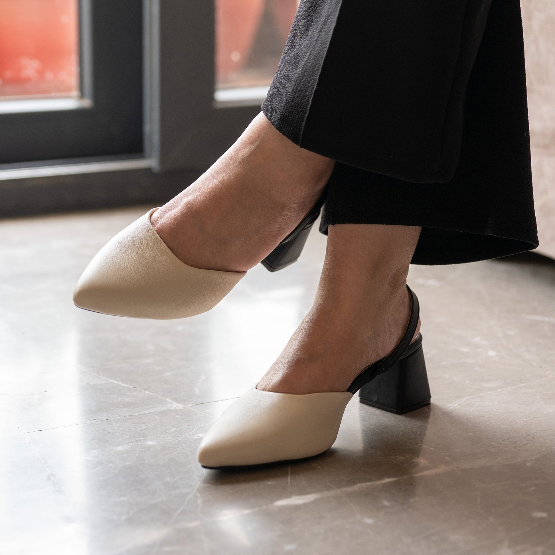 A pair of ivory pointed-toe Sandra block heels for women with a black block heel and sling back strap, worn with black pants, elegant heel shoes and high heel sandals for girls and women.